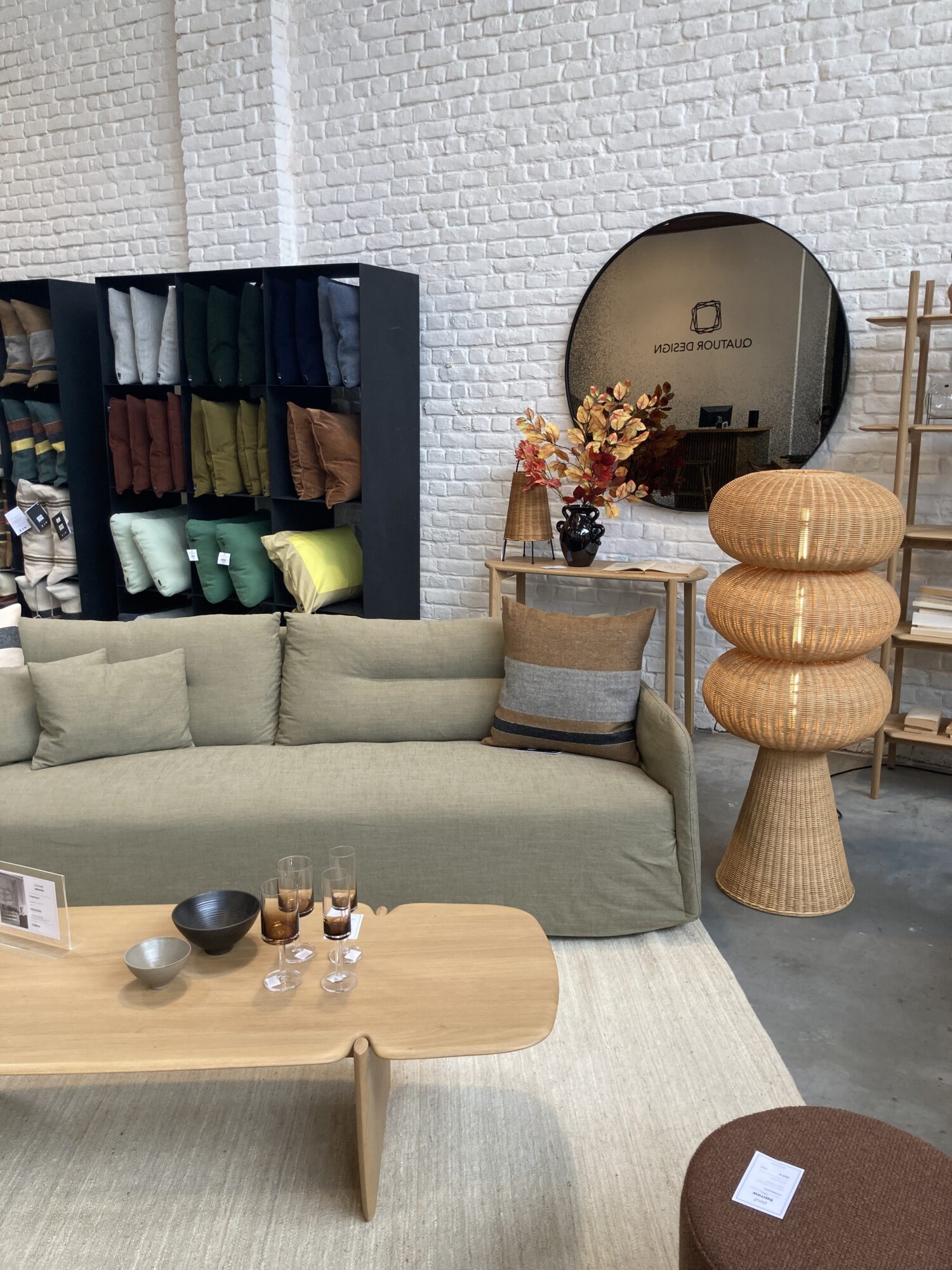 Un salon moderne provenant d'un Magasin meubles design Bruxelles, comprenant un canapé vert, une table basse en bois, une lampe tressée, un miroir rond, des vases décoratifs et des coussins soigneusement empilés sur des étagères contre un mur de briques blanches.