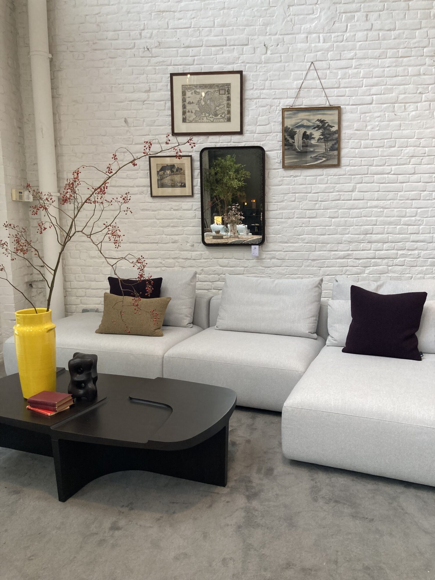 Un salon moderne avec un canapé sectionnel blanc, une table basse foncée, un vase jaune avec des branches et des œuvres d'art encadrées sur un mur de briques blanches - une inspiration parfaite d'un Magasin meubles design Bruxelles.