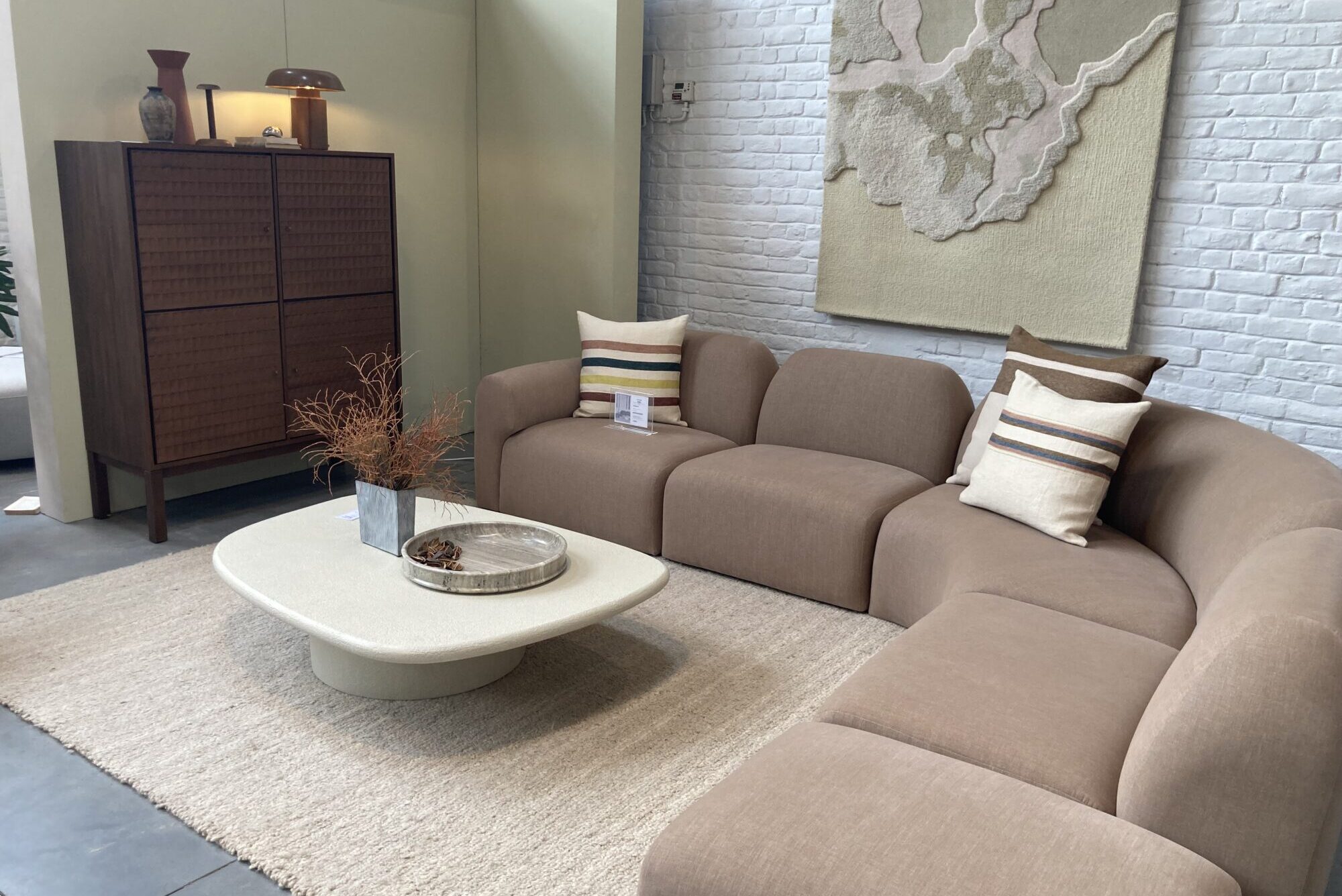 Un salon moderne conçu par quatuor design avec un canapé sectionnel beige, des coussins neutres, un tapis clair, une table basse, un meuble en bois et un art mural texturé sur un mur de briques blanches.