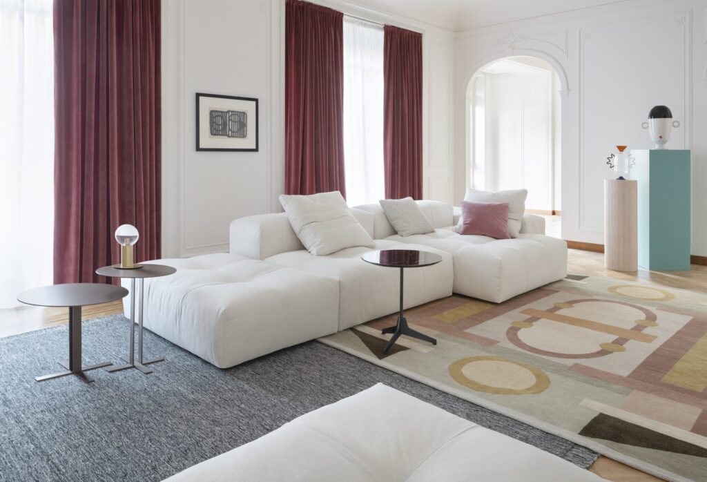 Salon moderne avec un canapé sectionnel blanc, deux petites tables rondes, des rideaux roses et un décor contemporain mettant en vedette le tapis Compo Pixel - Cat. C (2x1526, 1530, 3x1557, 1556, 3x1539, 2x1538) dans un cadre spacieux et lumineux.