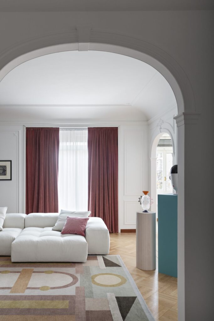 Un salon moderne avec un canapé sectionnel blanc, un tapis géométrique, des rideaux mauves et des objets décoratifs Compo Pixel - Cat. C (2x1526, 1530, 3x1557, 1556, 3x1539, 2x1538) sur un piédestal et une table d'appoint près d'une porte voûtée.