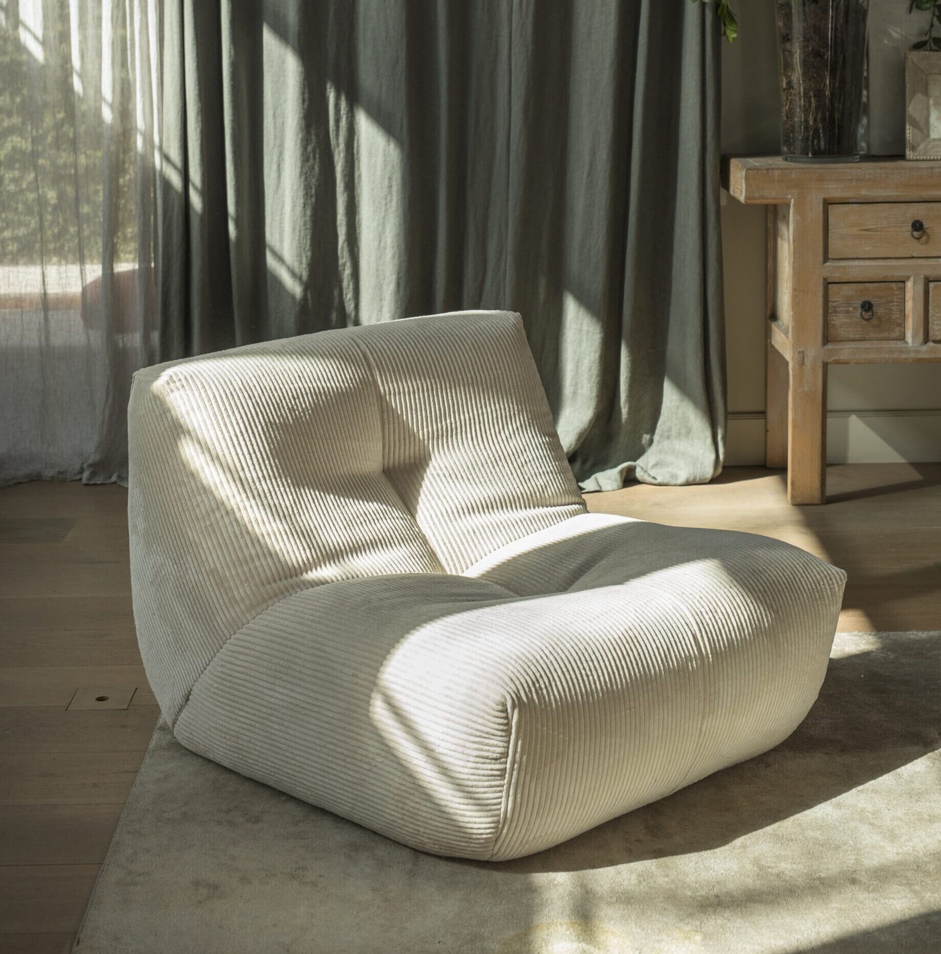 Une chaise longue en velours côtelé beige avec un dossier touffeté est posée sur un tapis clair dans une pièce ensoleillée avec un sol en bois, des rideaux et une table d'appoint en bois avec des plantes.