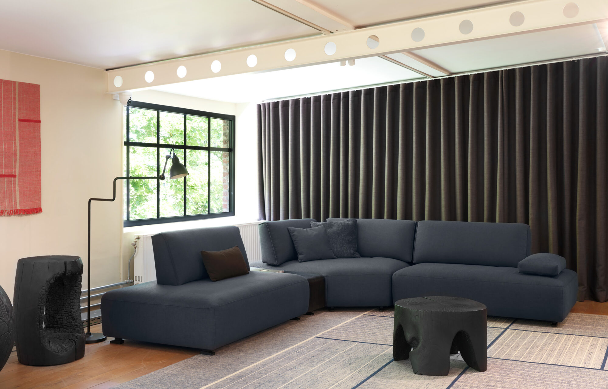 Un salon moderne avec un canapé sectionnel bleu foncé, des tables d'appoint noires, un lampadaire, une grande fenêtre et des rideaux noirs allant du sol au plafond.