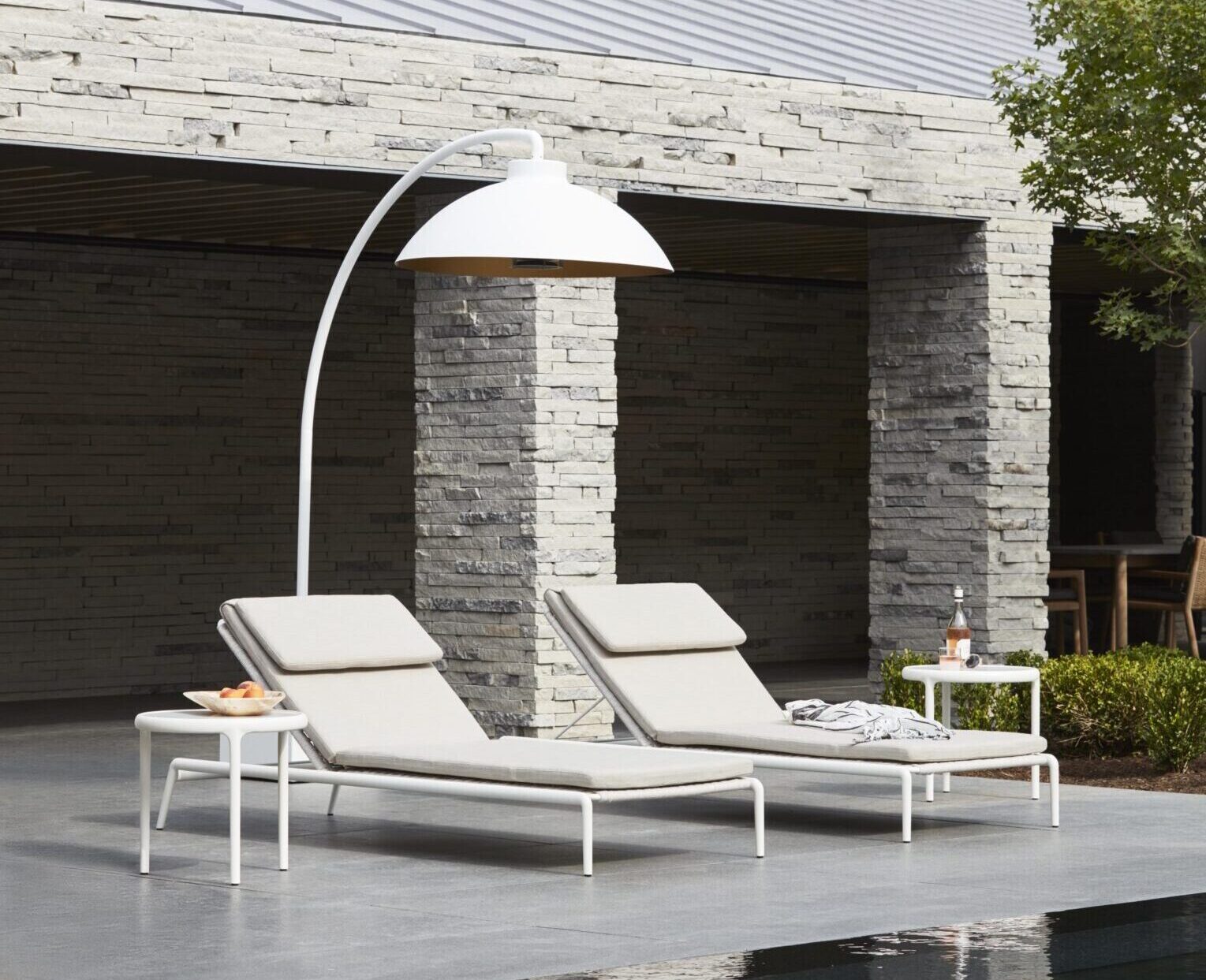 Deux chaises longues beiges et une table d'appoint avec des boissons et des snacks sont installées au bord d'une piscine, sous une grande lampe blanche, en face d'un bâtiment moderne en pierre.