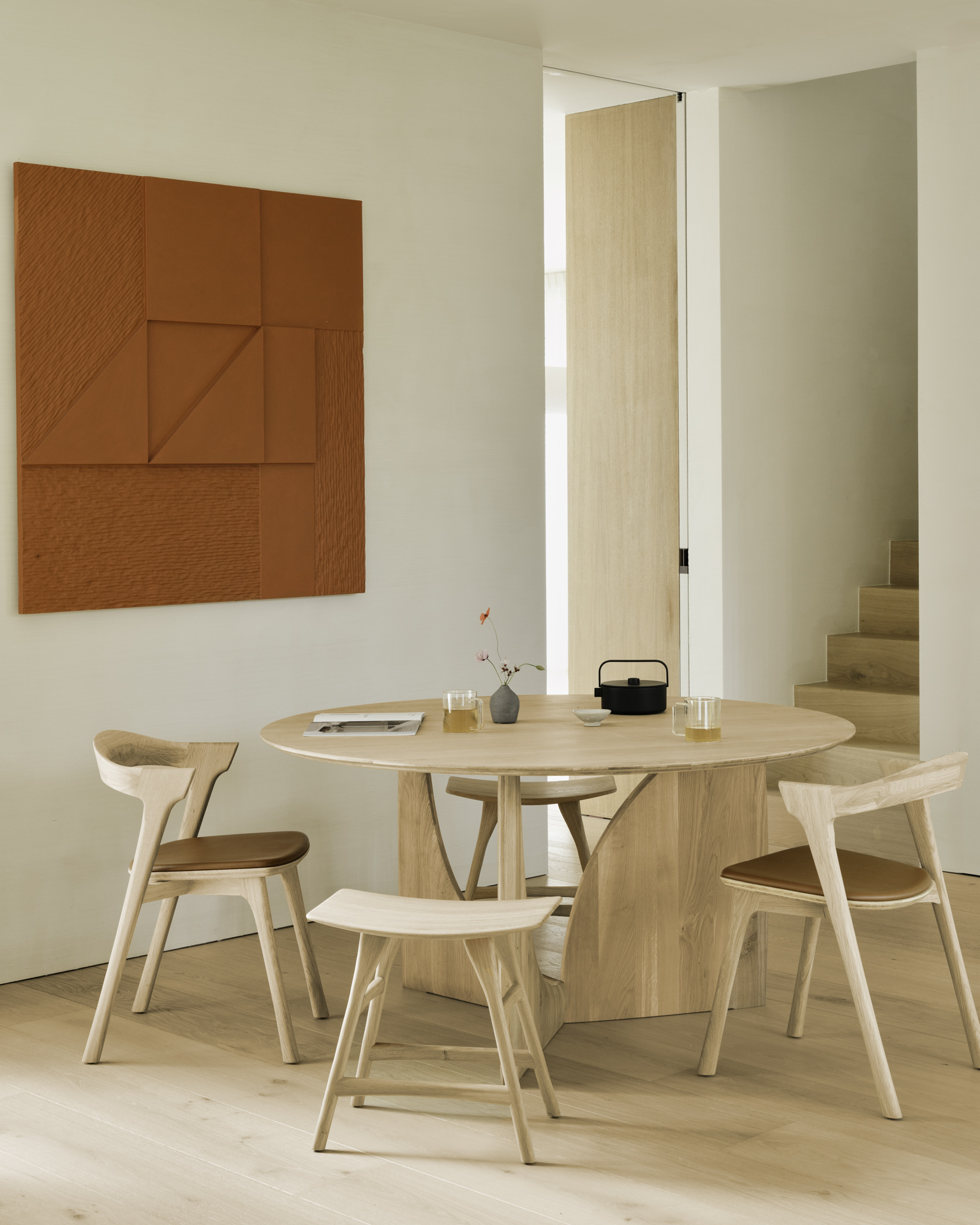 Une table ronde en bois avec trois chaises trône dans une pièce minimale et lumineuse ; une œuvre d'art géométrique orange est accrochée au mur et des escaliers sont visibles à l'arrière-plan.