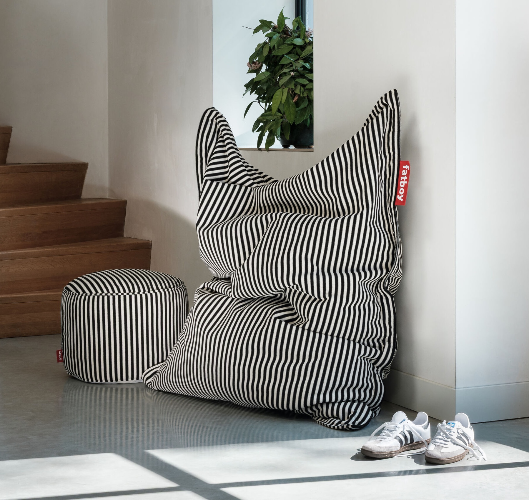 Un pouf rayé noir et blanc et un pouf assorti sont placés près des escaliers, avec une paire de baskets blanches et une plante en pot à proximité dans une pièce ensoleillée.