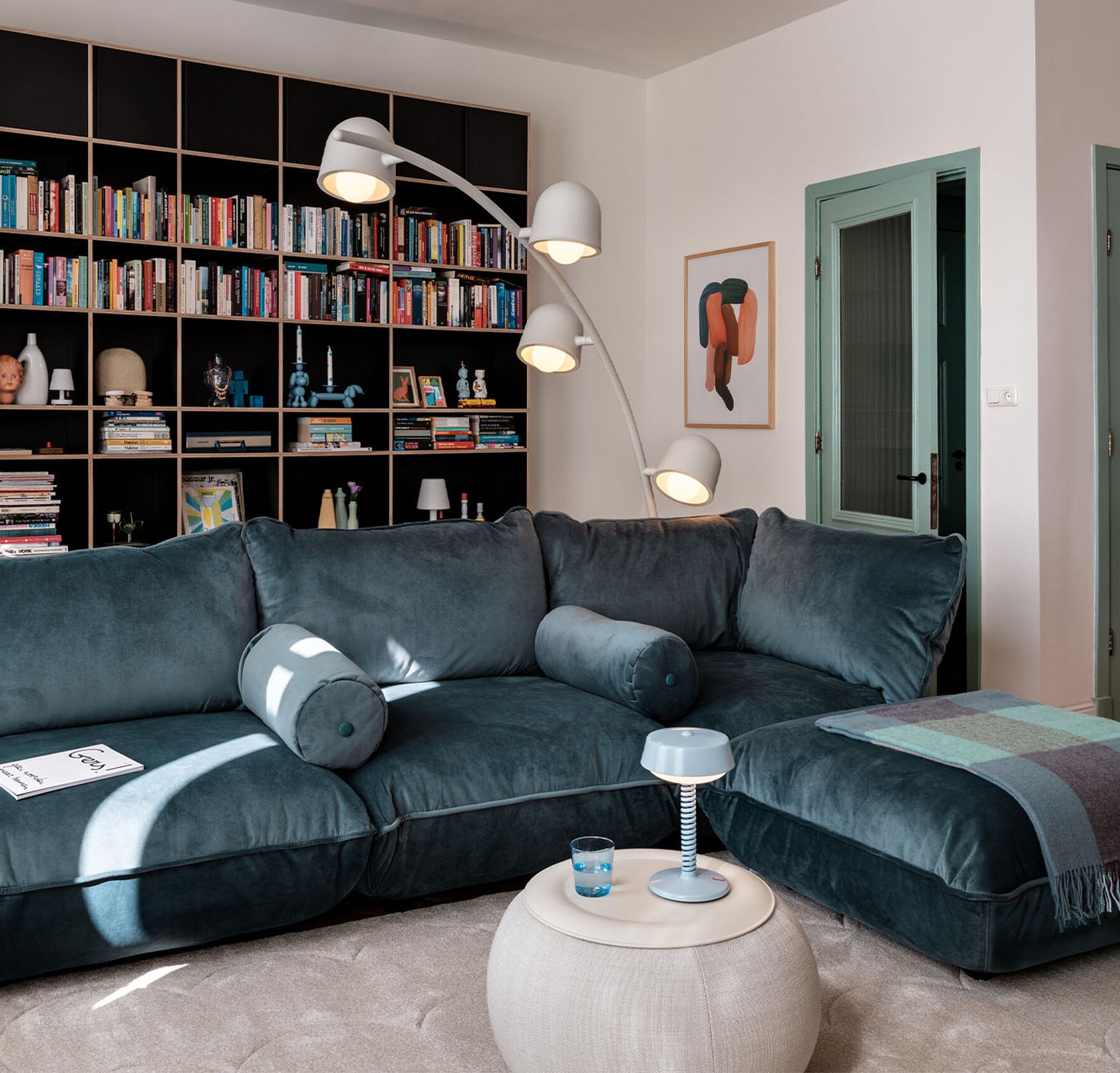 Un salon moderne avec un canapé sectionnel sarcelle foncé, une bibliothèque murale, un lampadaire en arc de cercle, un petit ottoman rond avec une lampe, et une couverture pliée sur le canapé.