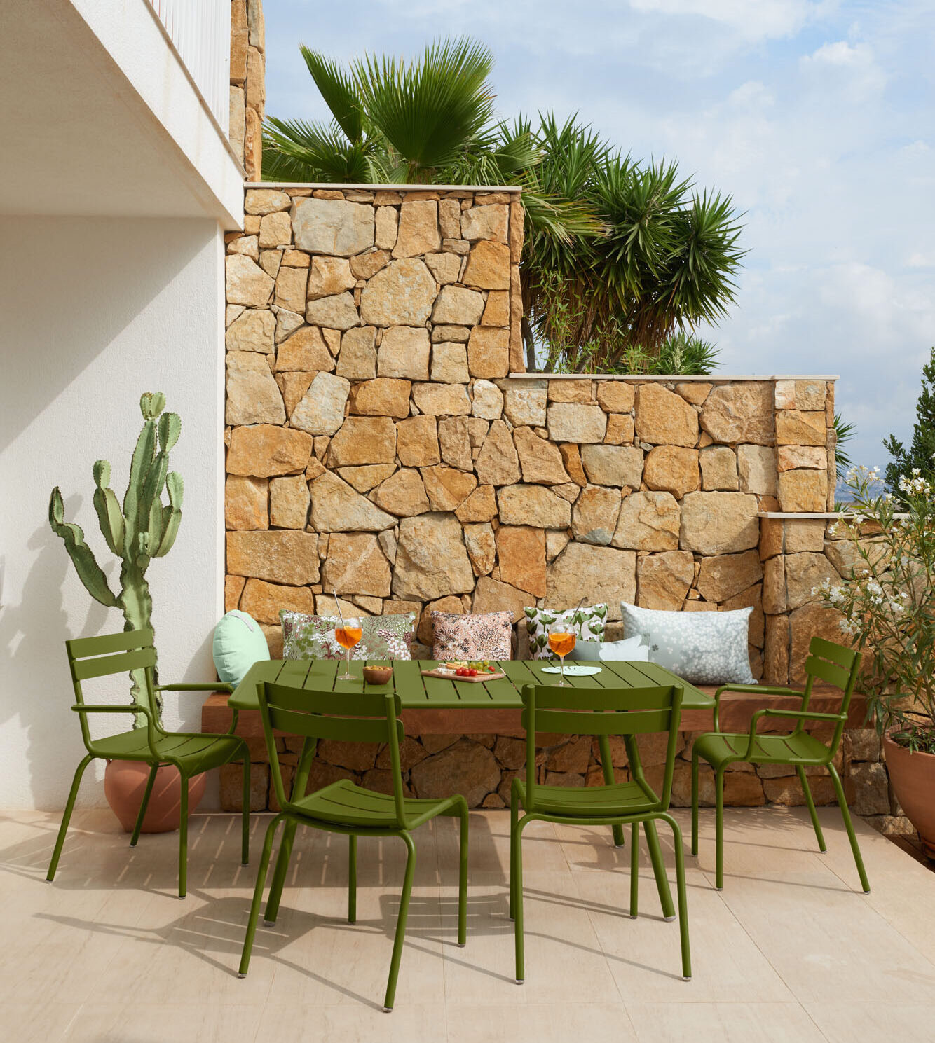 Une table de salle à manger verte et des chaises assorties sont disposées avec des aliments et des boissons sur un patio en pierre, entouré de plantes en pot et d'un mur en pierre.