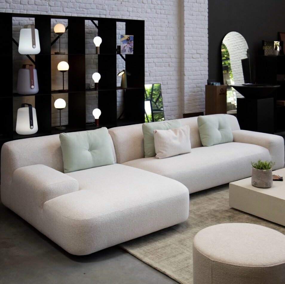 Un salon moderne avec un grand canapé sectionnel blanc, des coussins vert clair, un ottoman rond, une table basse et une étagère noire avec des lampes et des objets de décoration.