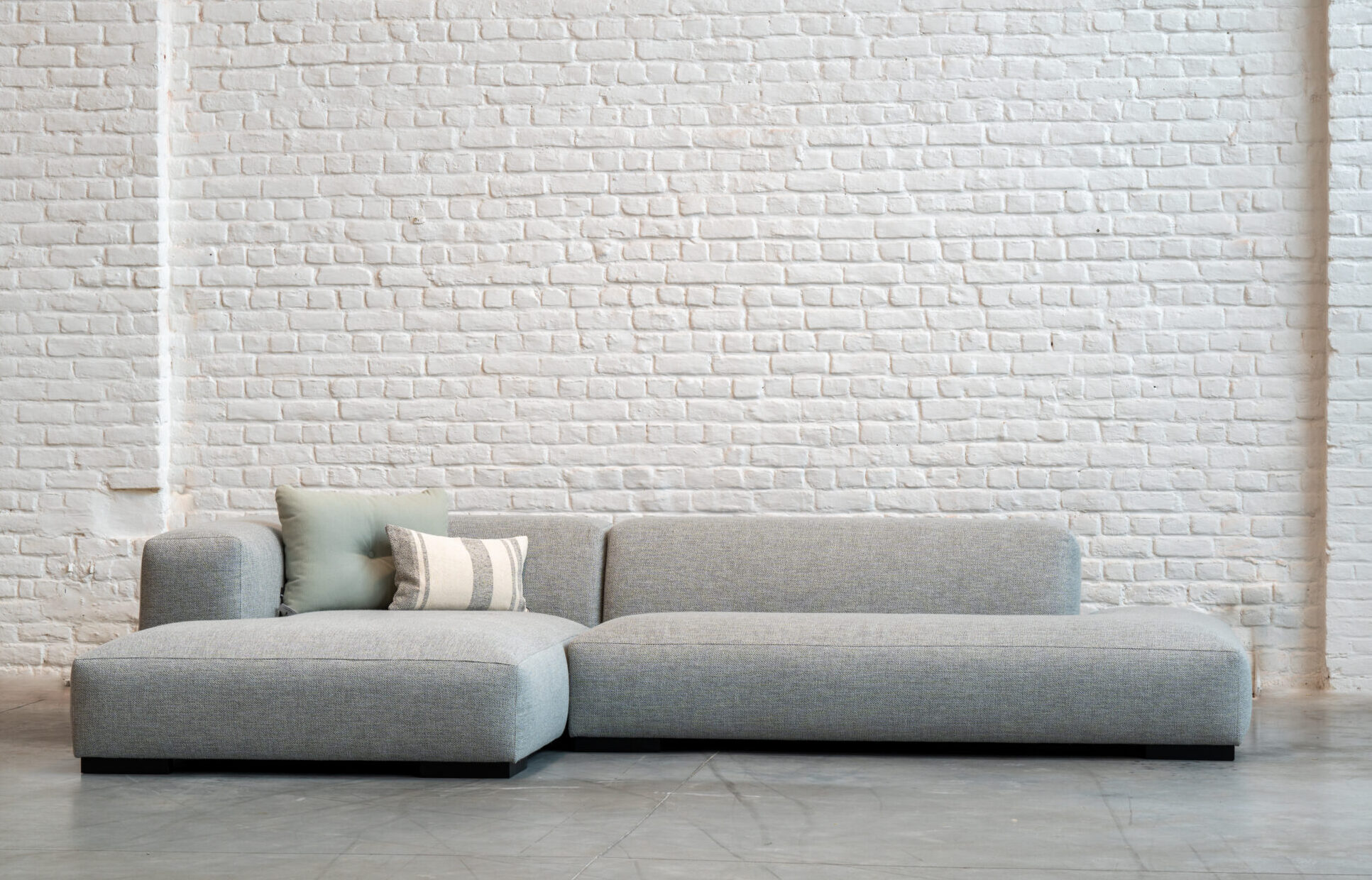 Un canapé sectionnel moderne gris clair avec deux coussins est placé devant un mur de briques blanches sur un sol en béton.