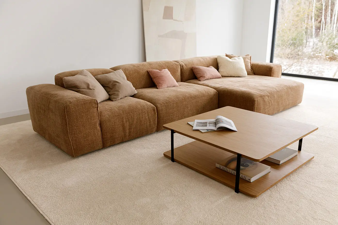 Un salon moderne avec un grand canapé brun, des coussins de couleur neutre, une table basse en bois avec des livres, et une grande fenêtre avec des arbres à l'extérieur.