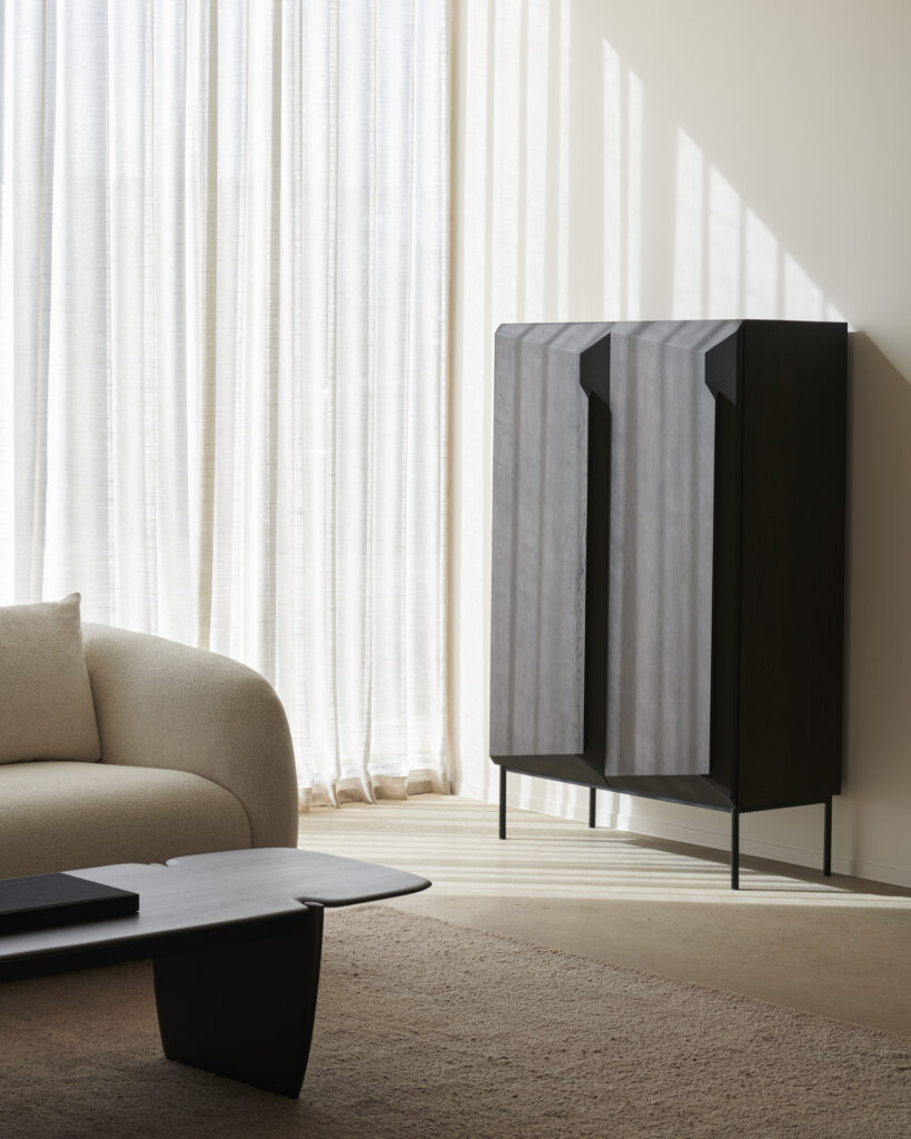 Un salon moderne avec un canapé beige incurvé, une table basse rectangulaire foncée et un grand meuble en chêne teinté brun près de rideaux blancs transparents.