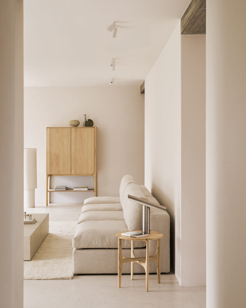 Salon minimaliste avec un canapé sectionnel beige, une armoire PI en chêne naturel huilé, une table basse et un décor neutre dans un espace doucement éclairé.