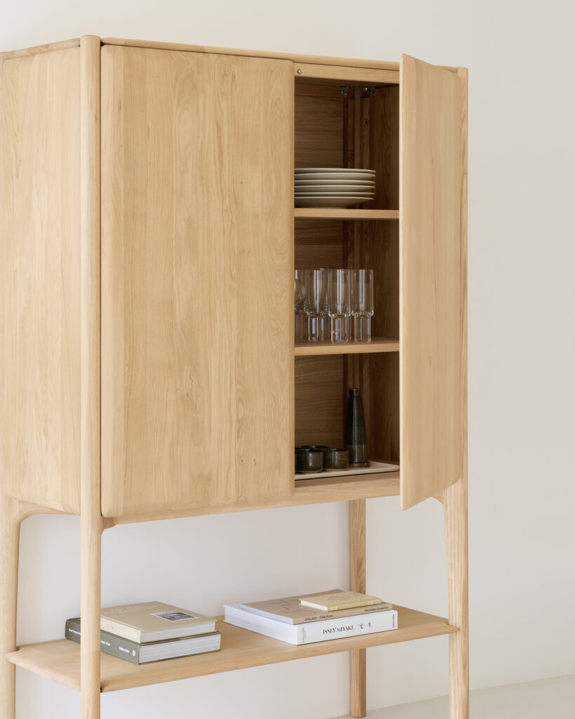 Une armoire PI en chêne naturel clair, dont une porte est ouverte, révèle les assiettes, les verres et les bouteilles à l'intérieur ; les livres et les magazines sont bien rangés sur l'étagère inférieure. Ce meuble huilé ajoute une touche de chaleur et d'élégance à tout espace.
