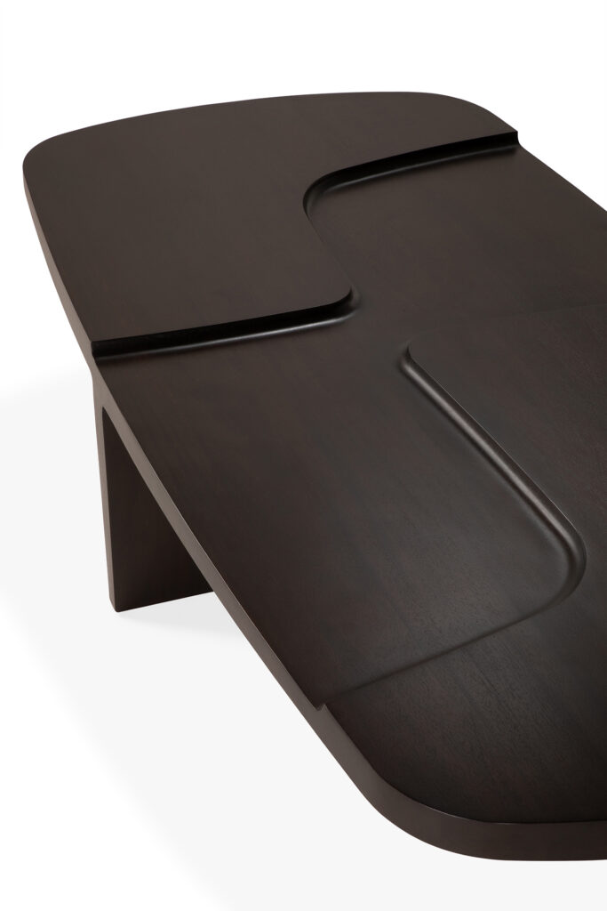 La table basse brutaliste est fabriquée en bois foncé avec des bords arrondis et des rainures asymétriques sculptées sur la surface, présentées sur un fond blanc.