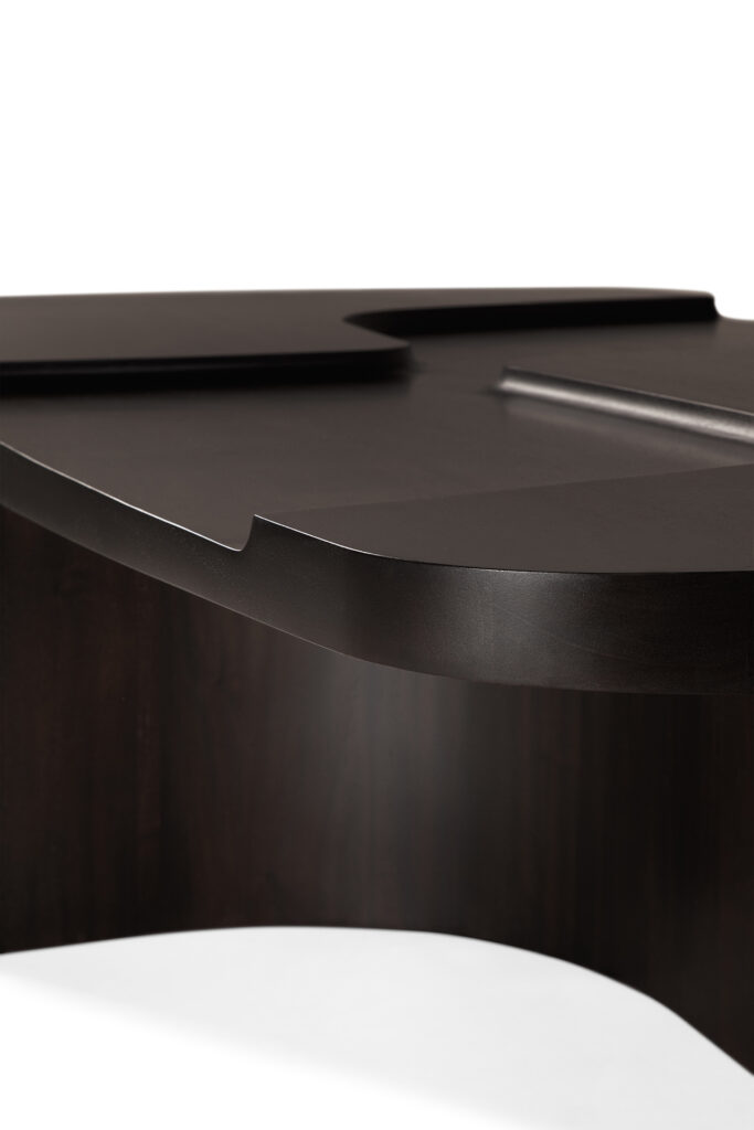 La table basse brutaliste se caractérise par une surface en bois sombre, un bord incurvé et une finition lisse. Elle est présentée en gros plan sur un fond blanc.