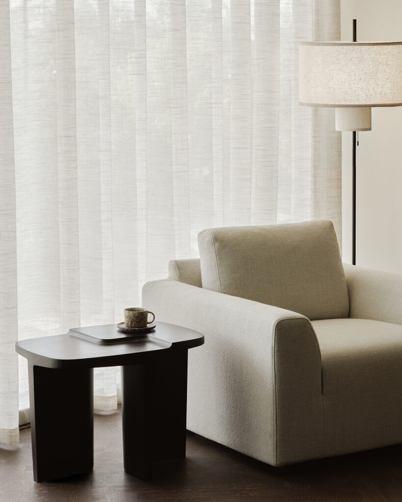 Un fauteuil beige est assis à côté de la table d'appoint brutaliste, qui contient une tasse et une soucoupe, à côté de rideaux transparents et d'un lampadaire dans un salon moderne.