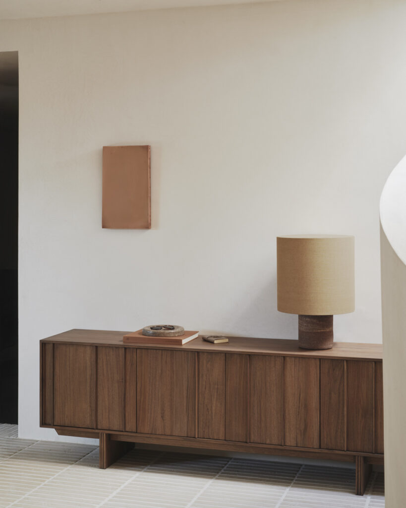 Un buffet pilier à 2 portes en teck teinté brun est posé contre un mur blanc uni, surmonté d'une lampe, de deux livres et d'un bol décoratif. Au-dessus, un petit tableau rectangulaire complète la scène minimaliste.