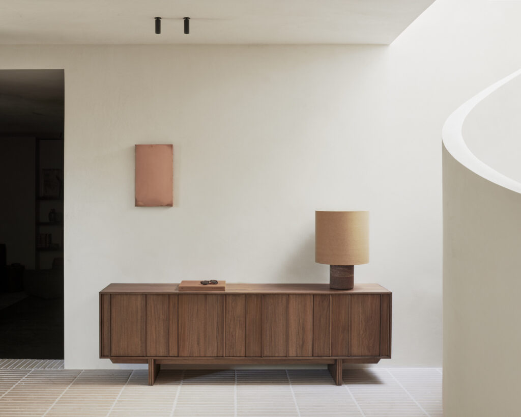 Un buffet pilier à 2 portes en teck teinté brun accueille une lampe beige et un petit plateau ; le décor minimal comprend une œuvre d'art murale rectangulaire exposée au-dessus sur le mur clair.