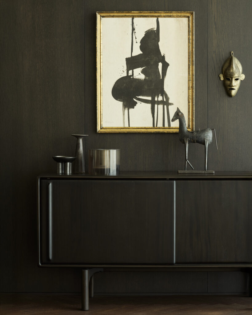 Un buffet Flow moderne avec des objets décoratifs, dont une sculpture de cheval en métal, un récipient en verre et un vase, est placé sous une peinture abstraite encadrée et un masque fixé sur un mur en bois d'acajou foncé.