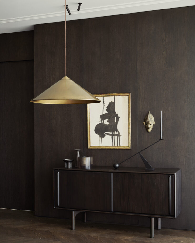 Une suspension en laiton est suspendue au-dessus d'un buffet en bois foncé acajou Flow, orné d'une décoration minimaliste, d'œuvres d'art et d'un masque africain accroché à un mur lambrissé de bois foncé.