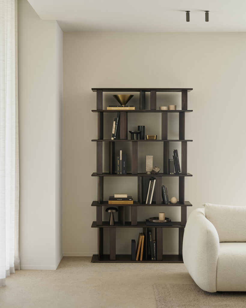 Une étagère en bois foncé, contenant des livres et des objets de décoration, est posée sur un mur beige à côté d'un canapé blanc dans un salon minimaliste.