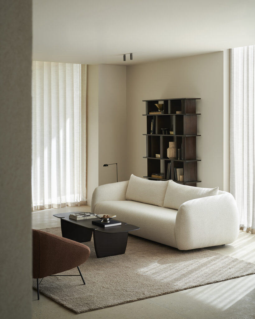 Salon minimaliste avec un canapé blanc, une table basse noire, un fauteuil marron, une étagère Block rack et de grandes fenêtres avec des rideaux transparents.
