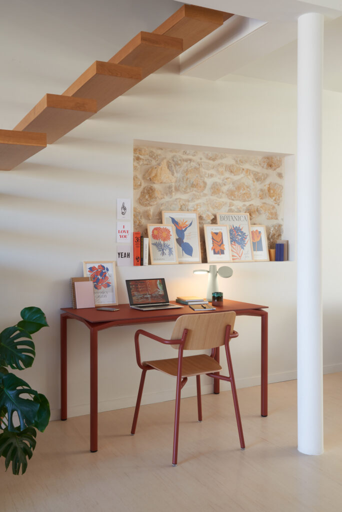 Un espace de travail moderne avec la Table Calvi en Velouté Ocre Rouge, associée à une chaise en bois, un ordinateur portable, une lampe et des œuvres d'art encadrées sous un escalier flottant en bois. Une plante verte est posée à côté du bureau.