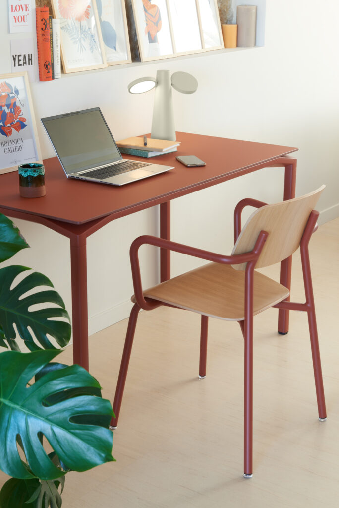Une table Calvi rouge avec un ordinateur portable, un cahier fermé, un téléphone, une lampe et des livres se trouve à côté d'une chaise en bois et d'une grande plante verte - un espace de travail accueillant dans les tons chauds de l'Ocre Rouge.