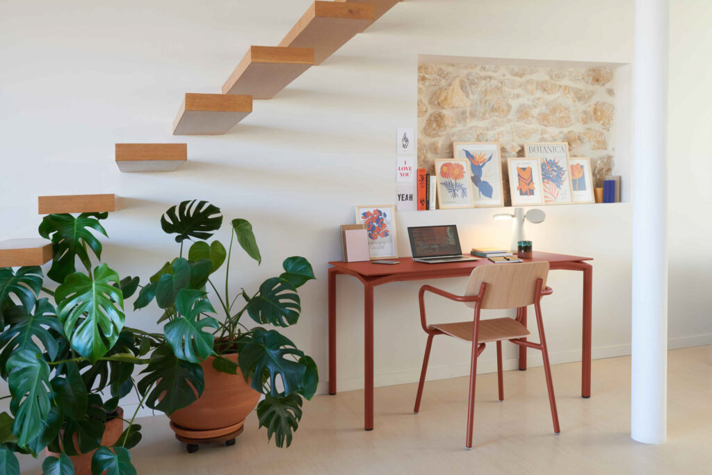 Un espace de travail minimaliste avec une Table Calvi en Velouté Ocre Rouge, une chaise en bois, un ordinateur portable, des livres, des œuvres d'art et une plante en pot, situé sous un escalier flottant en bois contre un mur blanc.