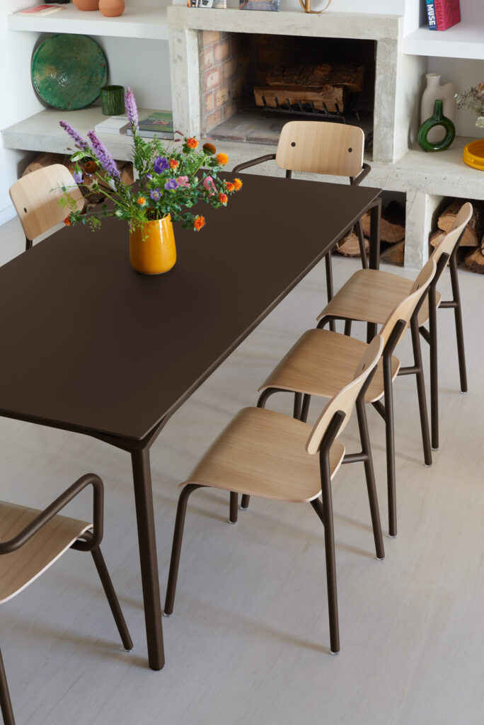 Une table Calvi 195x95 avec six chaises en bois est placée près d'une cheminée. Un vase jaune avec des fleurs assorties orne le centre, tandis que des étagères en Gris argile exposent des poteries et des livres à l'arrière-plan.