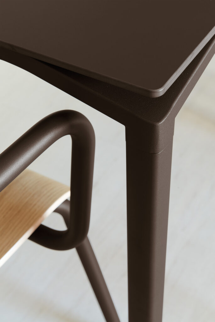 Gros plan d'une table Calvi 195x95 en gris argile, présentant un coin de table en métal brun et une chaise avec une assise en bois et une structure métallique incurvée sur un sol de couleur claire.