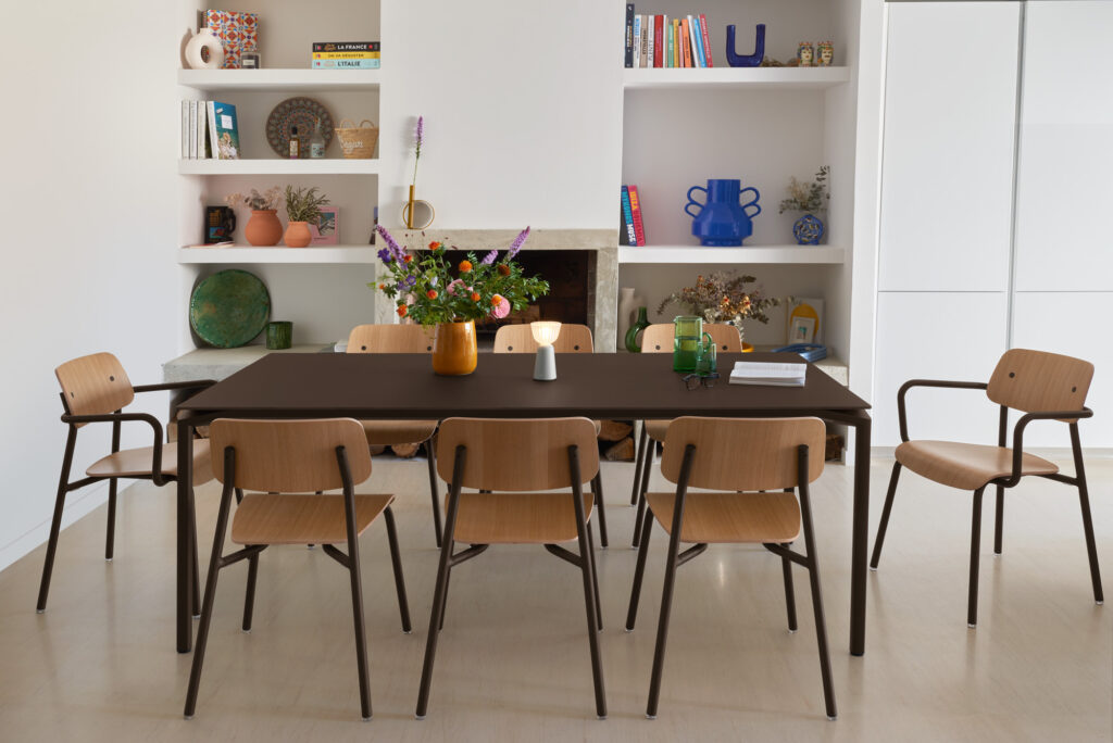 Une salle à manger avec une table Calvi 195x95 foncée, sept chaises en bois, un vase de fleurs et des étagères avec des livres, des céramiques et un décor ocre rouge contre un mur blanc.