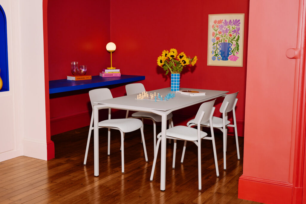 Une table Calvi (195x95) avec quatre chaises blanches est posée sur un sol en bois contre des murs ocre rouge. Un vase de tournesols, un jeu d'échecs et des fournitures artistiques sont posés sur la table ; des livres et des œuvres d'art se trouvent à l'arrière-plan.