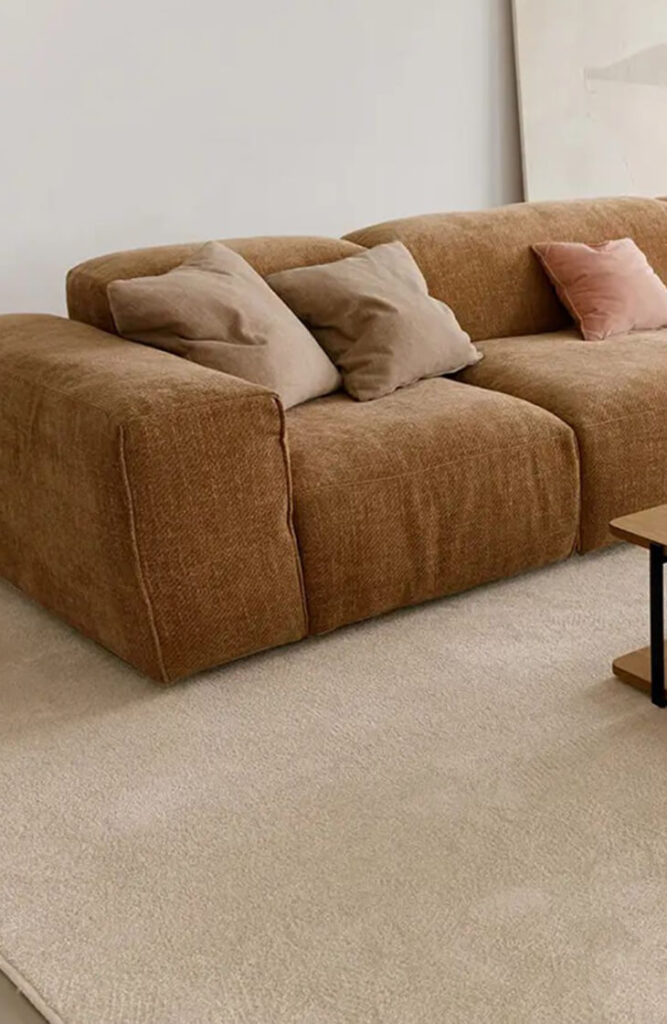 Un canapé sectionnel marron avec des coussins beiges et roses est posé sur un tapis clair à côté d'une table basse en bois dans cette pièce minimaliste, inspirée par le design quatuor d'un magasin de meubles design.