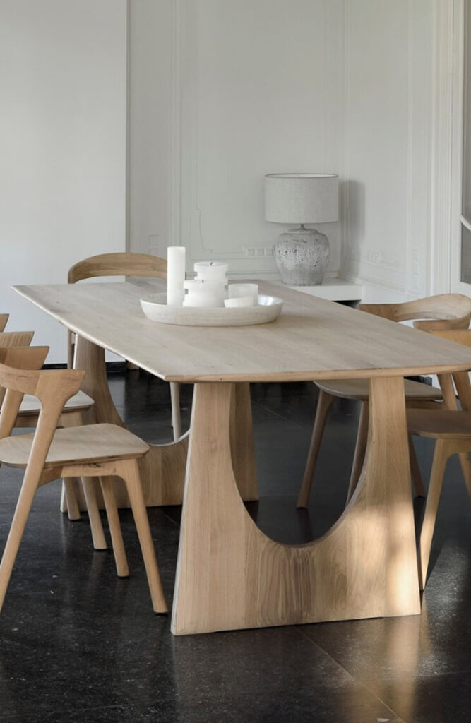 Une table de salle à manger moderne en bois clair de quatuor design est entourée de chaises assorties. La table accueille des bougies blanches et un plateau, tandis qu'une lampe en céramique est posée sur un buffet à l'arrière-plan.