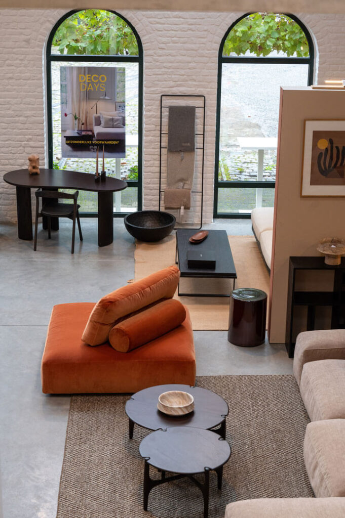 Salon moderne avec mobilier contemporain de quatuor design, comprenant un fauteuil orange brûlé, des tables rondes et un bureau noir près de fenêtres cintrées avec un poster "Deco Days".