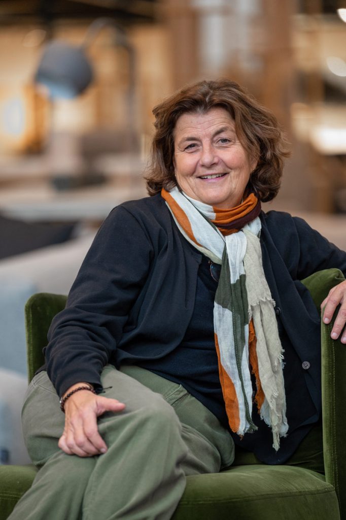 Une femme aux cheveux bruns ondulés, vêtue d'un haut noir, d'un pantalon vert et d'une écharpe multicolore, est assise avec assurance sur une chaise verte et sourit à la caméra, incarnant la chaleur et l'accessibilité des vrais professionnels.