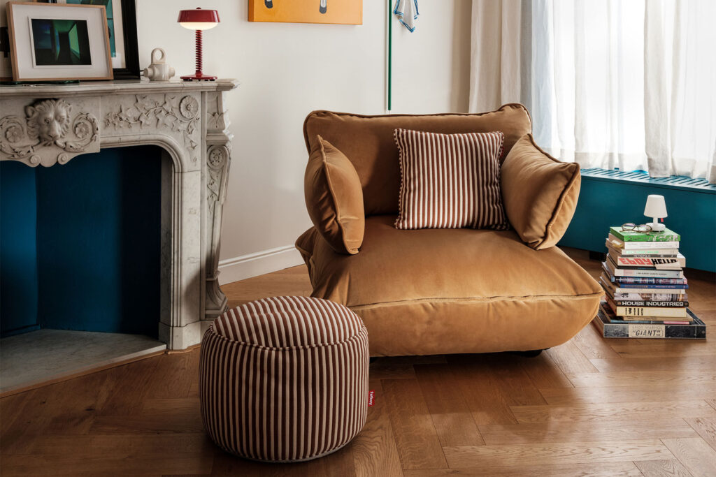 Le Sumo Loveseat, beige et spacieux avec un coussin et un ottoman rayés, se trouve à côté d'une cheminée ornée, d'une pile de livres et d'une fenêtre avec des rideaux diaphanes dans un salon confortable.