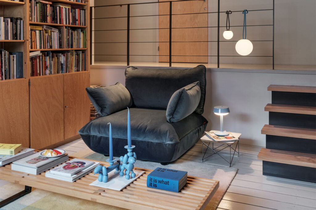 Créez un coin lecture confortable avec le grand canapé Sumo en gris foncé, des étagères, une table basse en bois avec des livres et des bougies, ainsi que des lampes suspendues et des lampes de table modernes et élégantes.