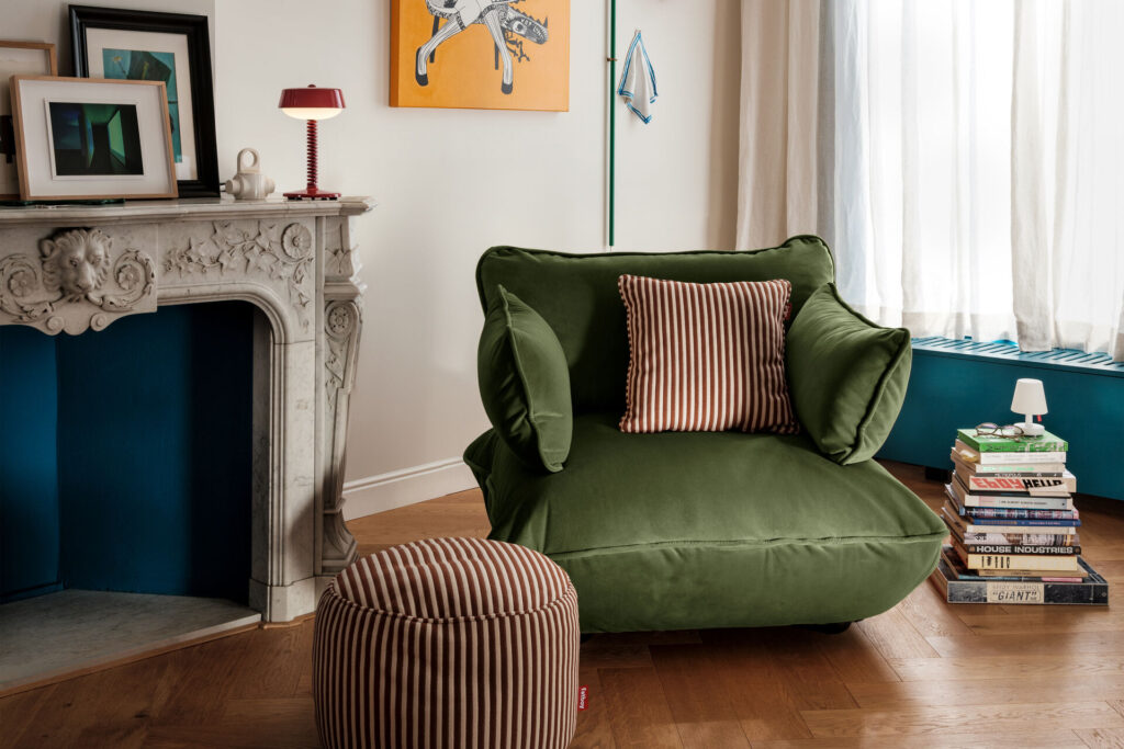 La Causeuse Sumo en vert, avec des coussins rayés et un ottoman assorti, est placée près d'une cheminée en marbre, avec des livres et une petite lampe, dans un salon moderne et confortable.