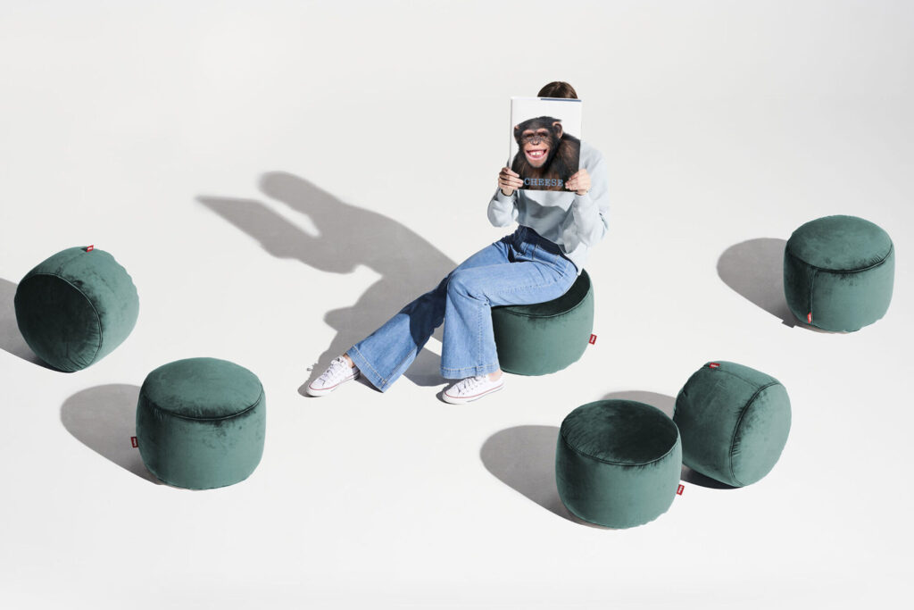 Une personne est assise sur un pouf Point Velvet Recyclé - Petrol, tenant une photo de visage de chimpanzé, entourée de cinq ottomans recyclés assortis, le tout sur un fond blanc impeccable.