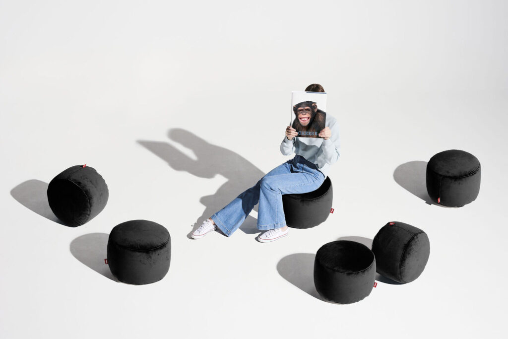 Une personne est assise sur un pouf Point Velvet Recycled - Petrol, tenant un magazine avec un visage de chimpanzé aligné sur sa propre tête, sur un fond blanc uni.