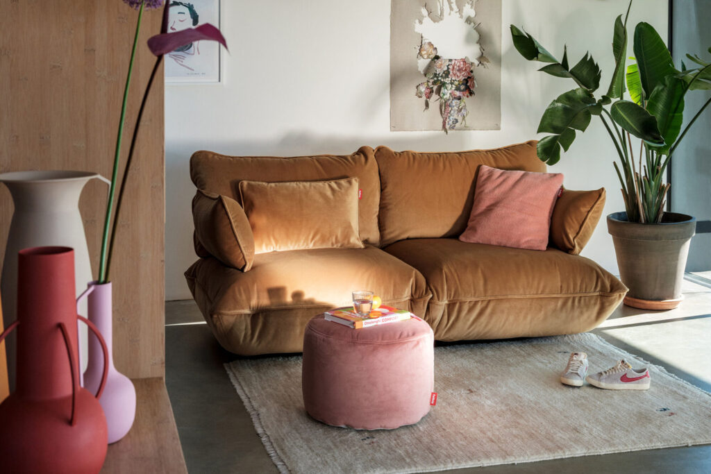 Un canapé beige avec des coussins Point Velvet Recycled-Petrol et beige est posé sur un tapis clair à côté d'un ottoman pétrole, de plantes, d'œuvres d'art murales, de vases et d'une chaussure de sport au sol.