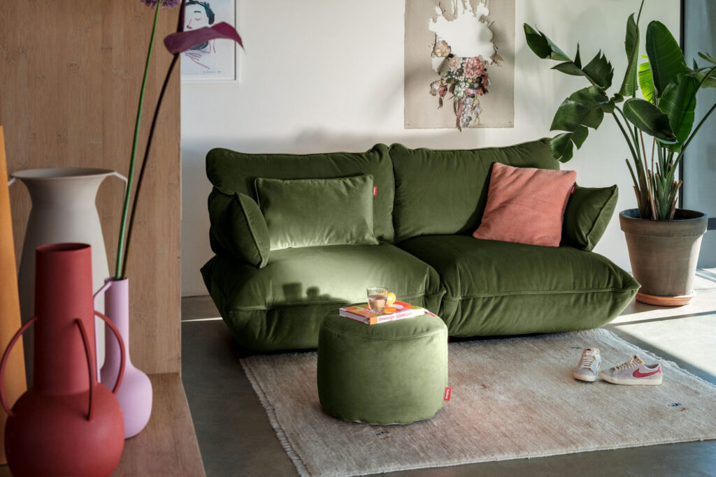 Un salon moderne présente le canapé Point Velvet Recycled - Petrol avec un coussin rose et un ottoman sur un tapis clair près d'une plante en pot ; des baskets rouges et blanches et des vases décoratifs sont également visibles.