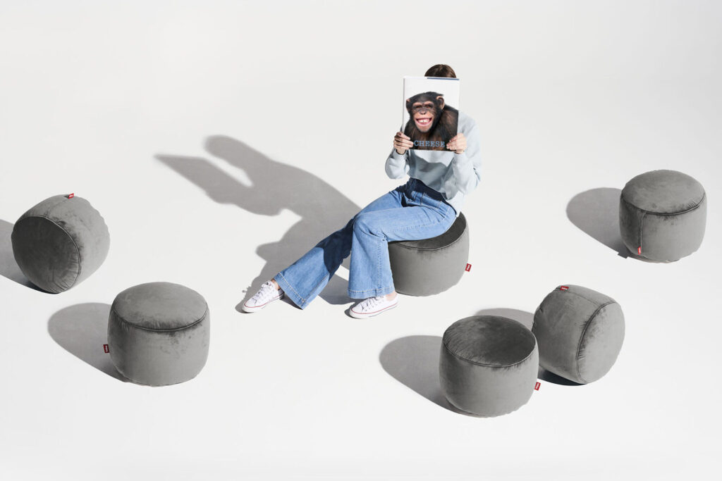 Une personne est assise sur un pouf Point Velvet Recyclé - Petrol, tenant une photo de chimpanzé devant son visage, entourée de cinq poufs recyclés similaires sur un fond blanc.