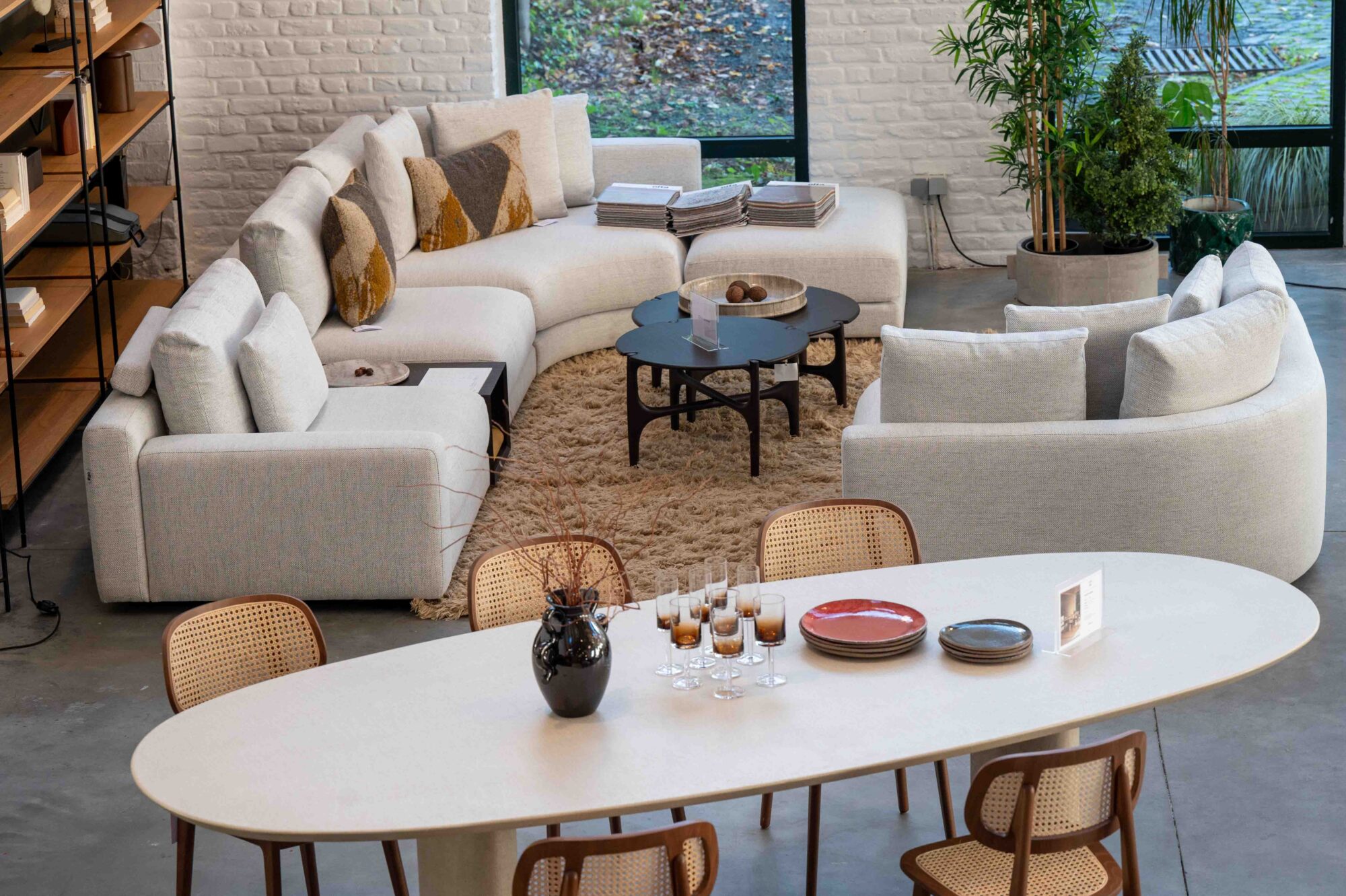 Salon et salle à manger modernes conçus pour les professionnels, comprenant un canapé Olta Stone modulable blanc incurvé, des tables basses rondes noires, une table de salle à manger ARI de V. Sheppard avec des chaises, et de grandes fenêtres mettant en valeur la verdure à l'extérieur.