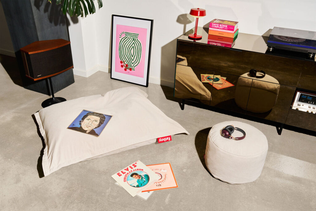 Un salon moderne avec un coussin de sol Fatboy Point Cord Deep Blue, des disques vinyles, des écouteurs, un pouf, des œuvres d'art encadrées, un haut-parleur et une crédence présentant une lampe, des livres et un tourne-disque.