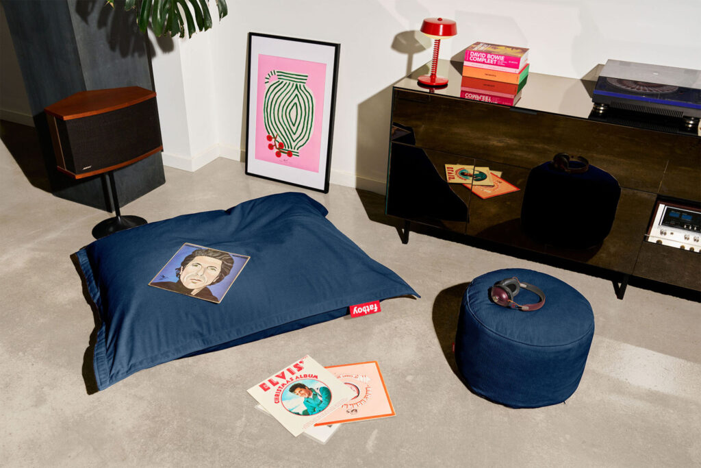 Un salon moderne avec un coussin de sol Fatboy Point Cord Deep Blue, des disques vinyles, un tourne-disque, un haut-parleur, un tableau encadré, un ottoman assorti et une table d'appoint avec lampe ajoute un confort élégant à votre espace.