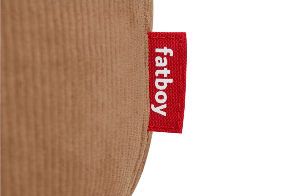 Gros plan sur une étiquette en tissu Fatboy rouge avec un texte blanc, attachée à une surface brune texturée. Cette étiquette fait partie de la collection confortable Fatboy Point Cord Deep Blue.